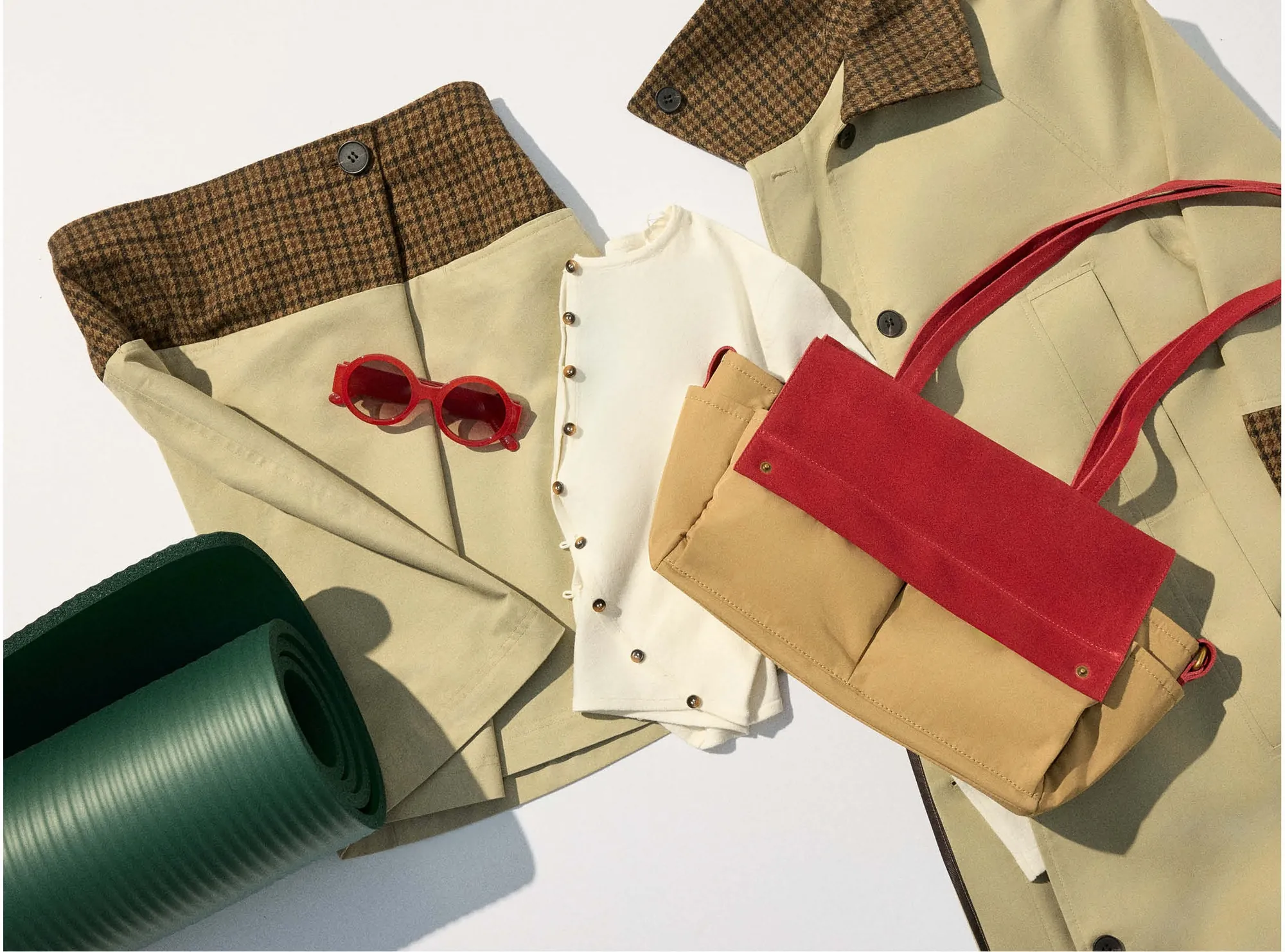 Flat lay: the trench coat, red shoulder bag, sunglasses and a rolled green mat