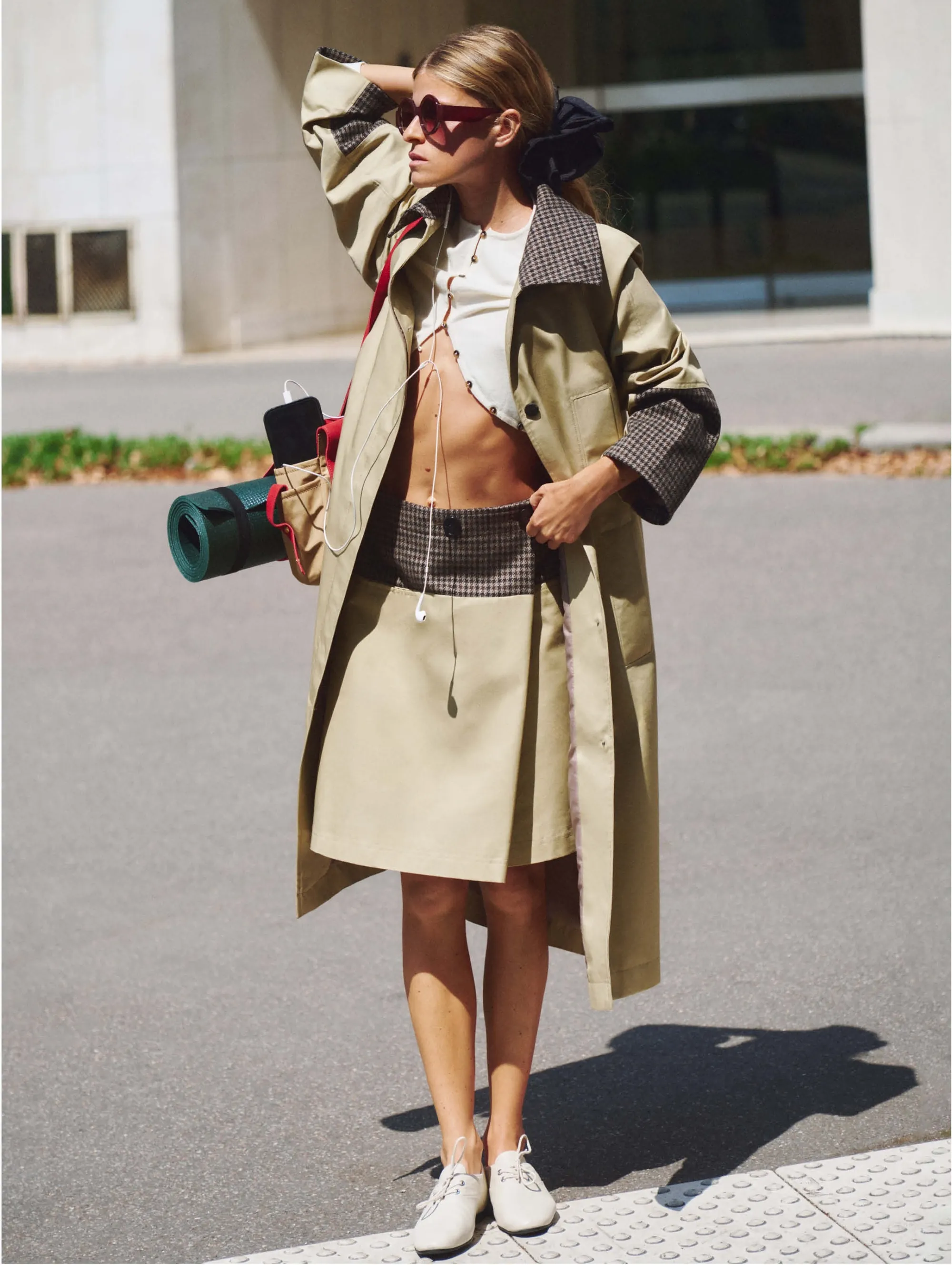 In the trench coat and camel skort on a sunny street, the mat bag over the shoulder