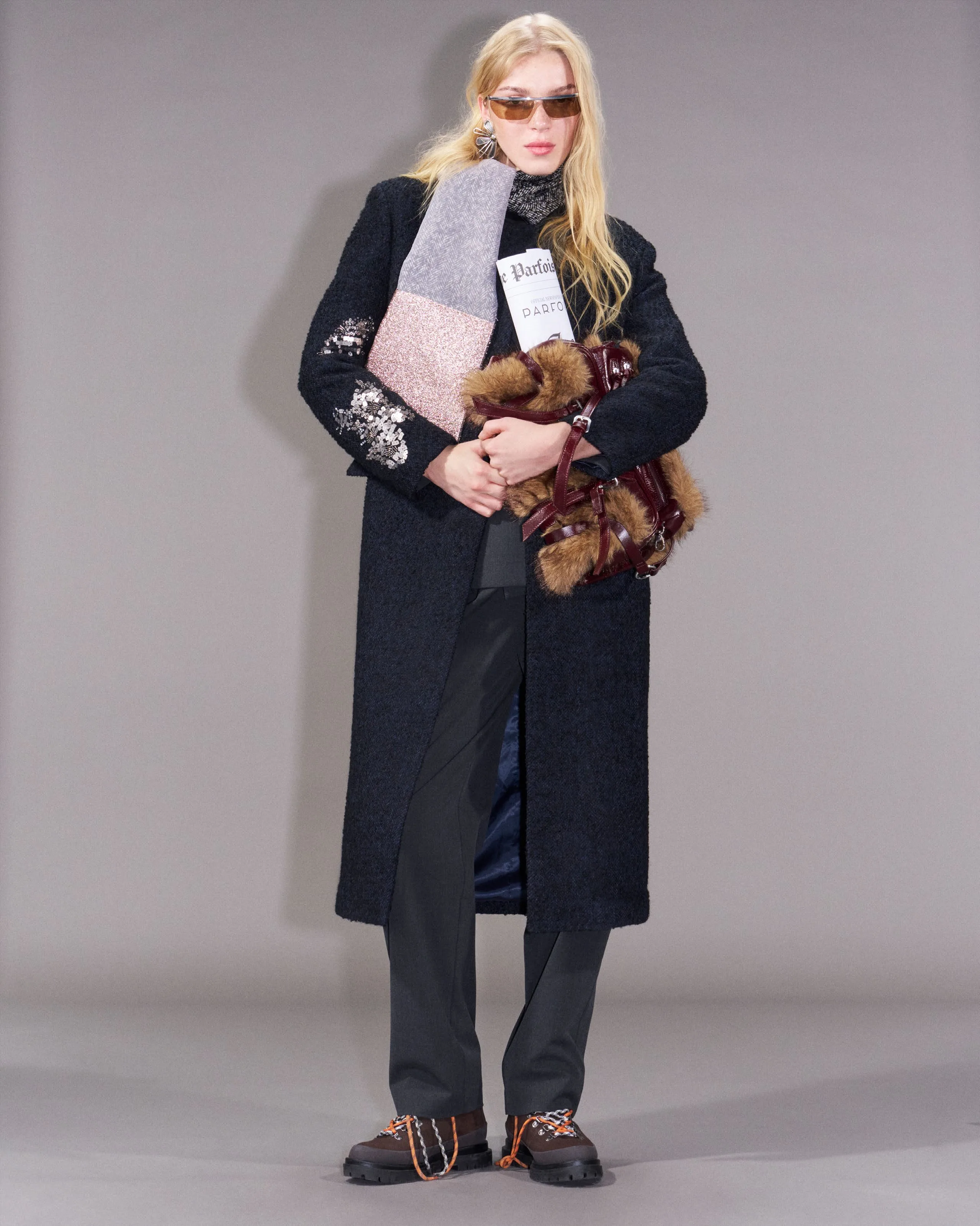 Colin in a long dark bouclé coat holding a fur bag and a newspaper
